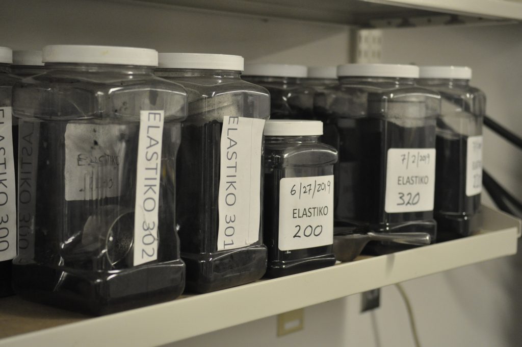 Plastic jars on a shelf in a lab containing different types of pavement mixtures.