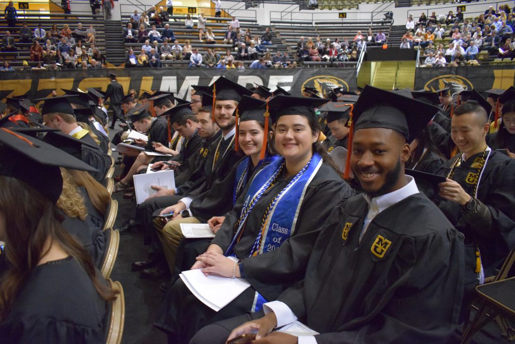 Mizzou Engineering graduates in their regalia smile at the camera from their seats in the Hearnes Center