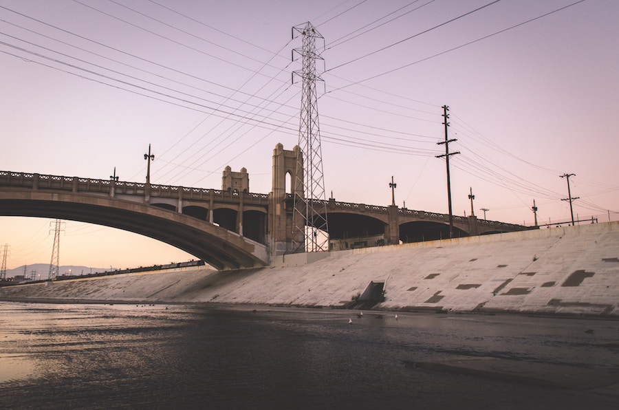 A big industrial bridge arching over a canal. Power lines criss-cross over a sky that deepens from tangerine to lavender.