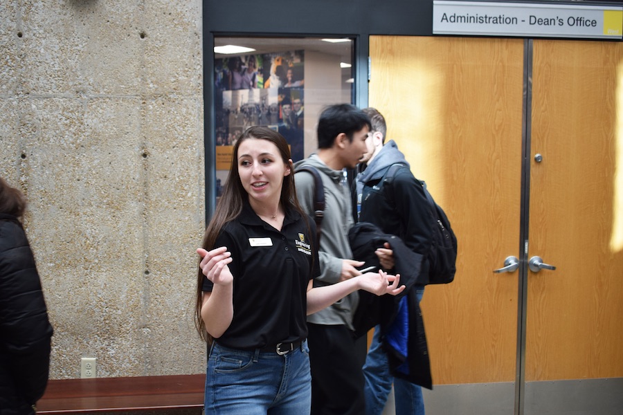 A young woman in a black polo with a name tag shows students around.