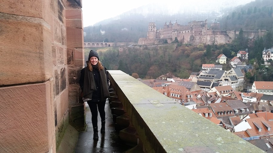Carly Garrow stands on a stone balcony. Behind her, the red roofs of Heidelberg, Germany, and halfway up the mountain is the Heidelberger Schloss.