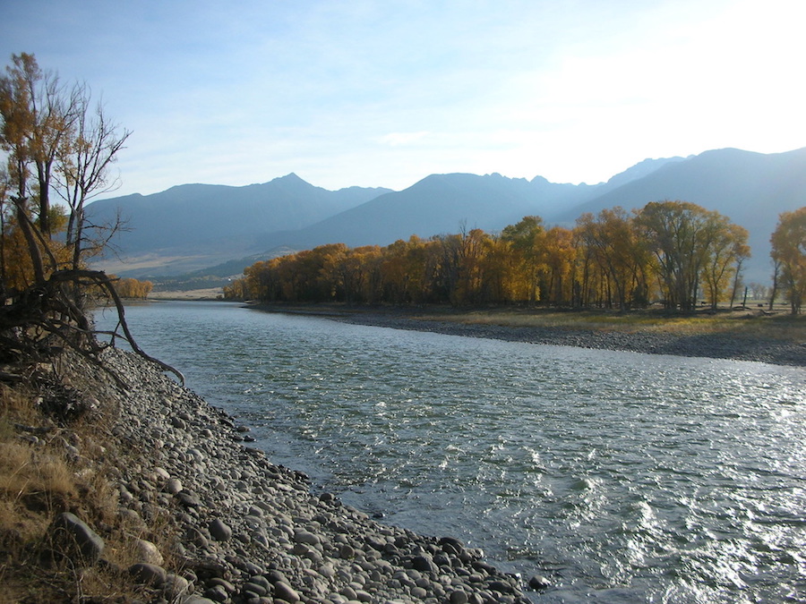 A landscape showing a narrow river shining under hazy sunlight, low mountains in the background, and trees on either side of the bank, dressed in autumn foliage.