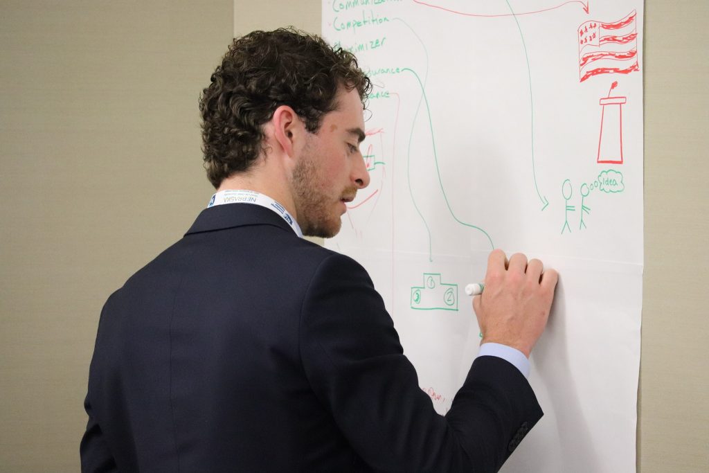 A young man in a suit writes on a markerboard.