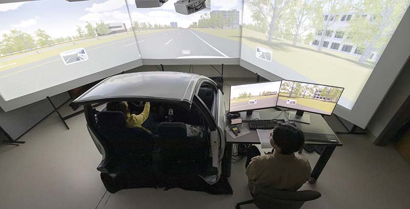 A woman sits in the front seat of a car cab surrounded by a simulated environment.