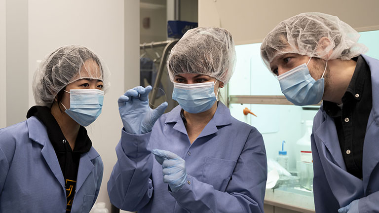 Three people in clean gear and PPE look at a specimen