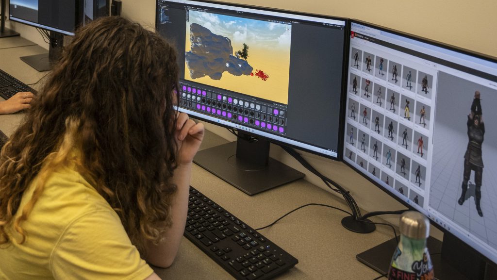 A young woman peers at computer game environment and avatars on dual display monitors