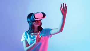 A young woman wears a virtual reality headset, actively reaching out with her hands as she explores a digital environment in a vibrant setting illuminated by colorful light.