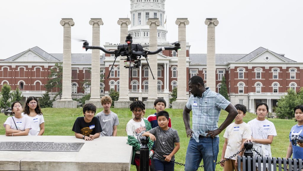 A group of middle-school students and an instructor watch a hovering quad copter