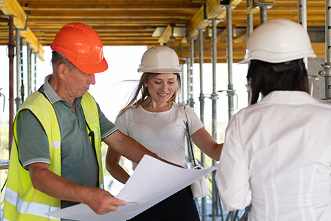 Three people in hard hats look at plans on a construction site