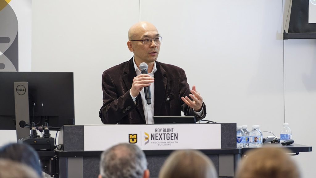 Jianlin Cheng speaks into a microphone at a podium with an audience seated in front.