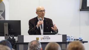 Jianlin Cheng speaks into a microphone at a podium with an audience seated in front.