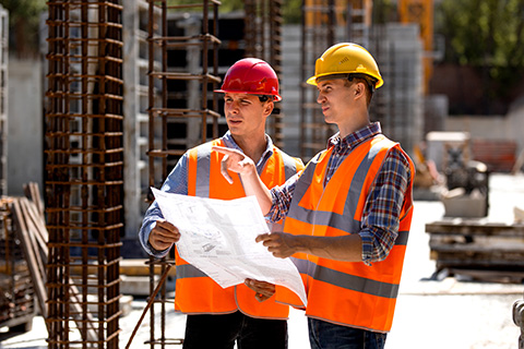 Construction manager and engineer dressed in orange work vests and hard helmets explore construction documentation on the building site near the steel frames